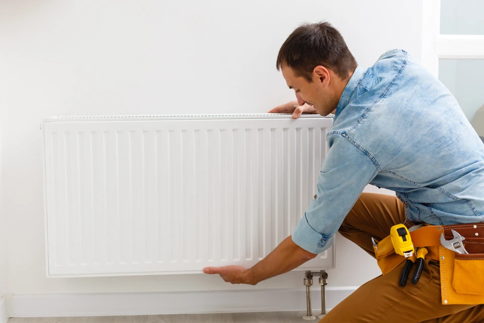 Un technicien en salopette beige et chemise en jean installe ou r&eacute;pare un radiateur &agrave; panneaux blanc dans un int&eacute;rieur r&eacute;sidentiel moderne