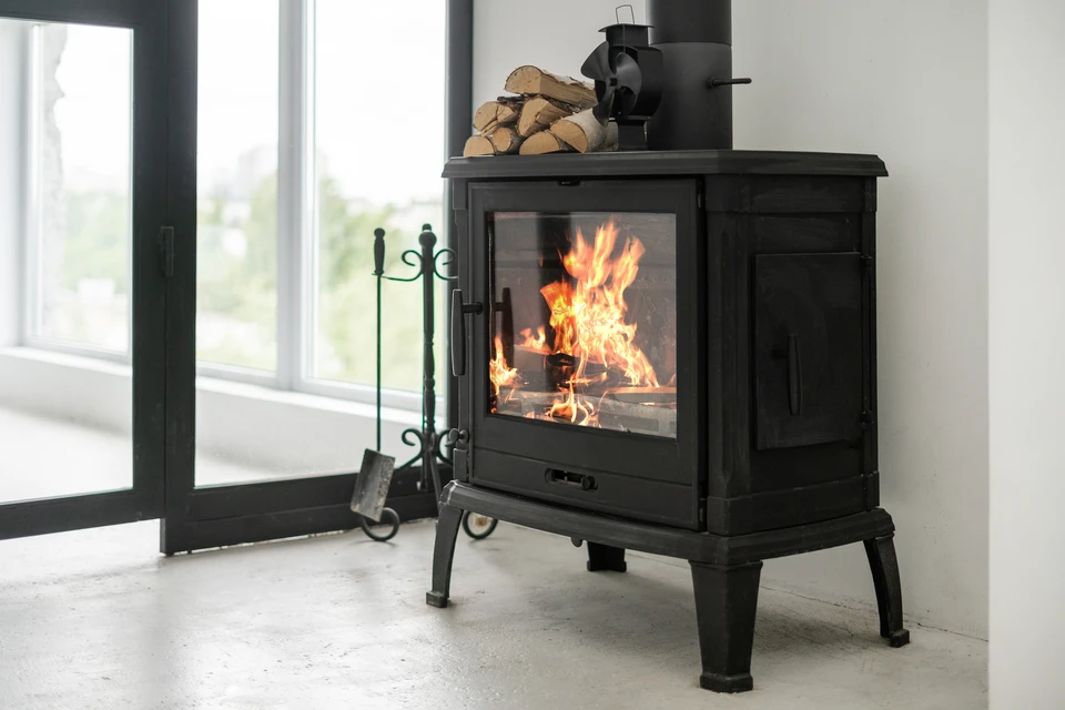 Intérieur chaleureux avec poêle à bois en fonte noire et porte vitrée, panier en osier contenant petit bois d'allumage, casier métallique rempli de bûches fendues